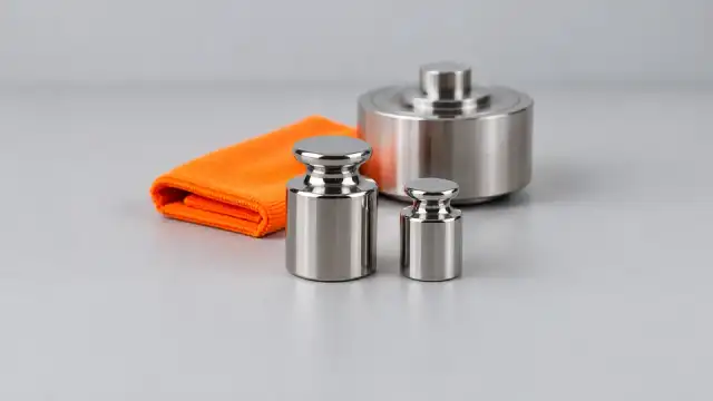 Pesos-padrão de inox e uma célula de carga de aço sobre a bancada, com um pano laranja dobrado